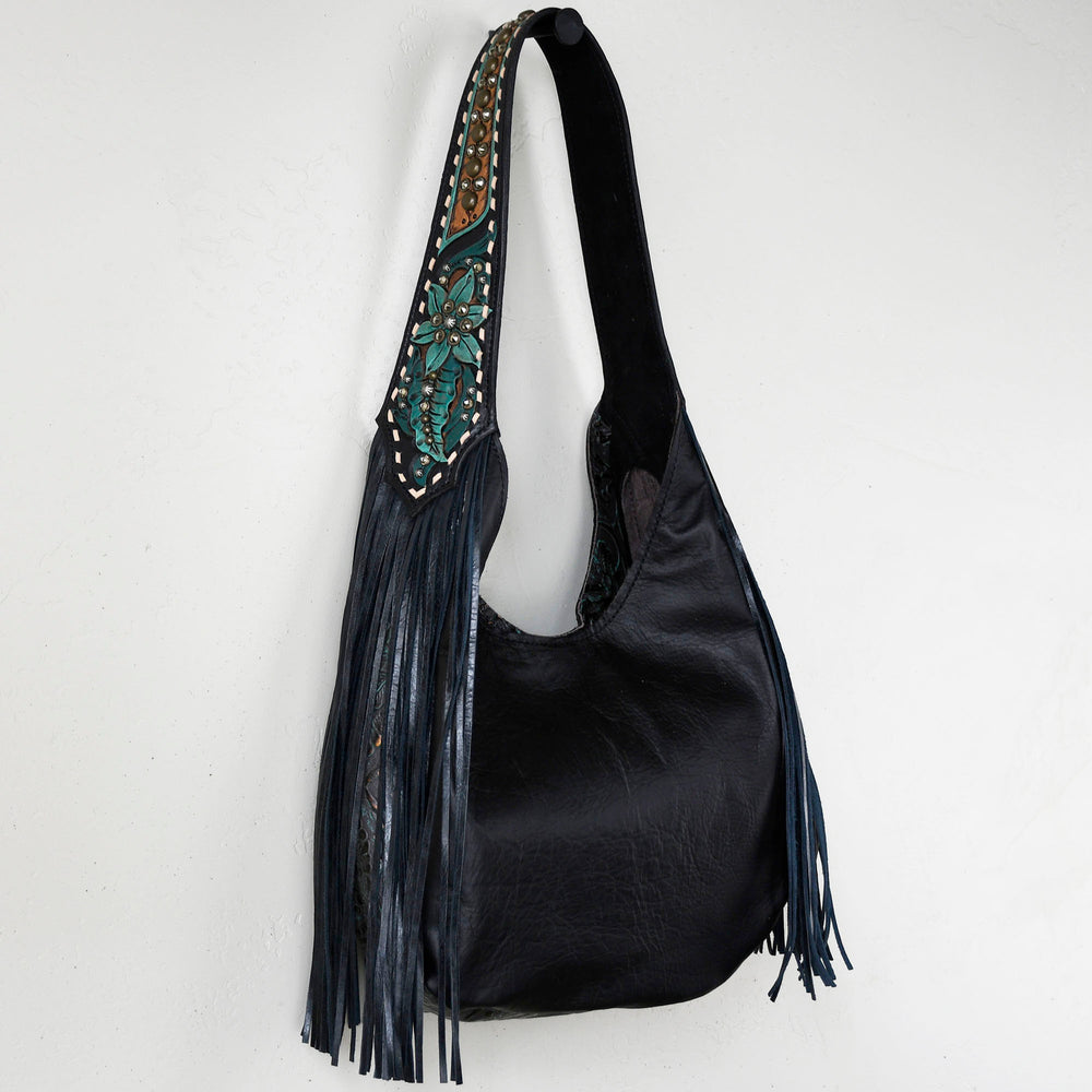 
                  
                    A black leather Marilyn bag #1 with fringe and a patterned strap hanging on a white wall by Heritage Brand.
                  
                