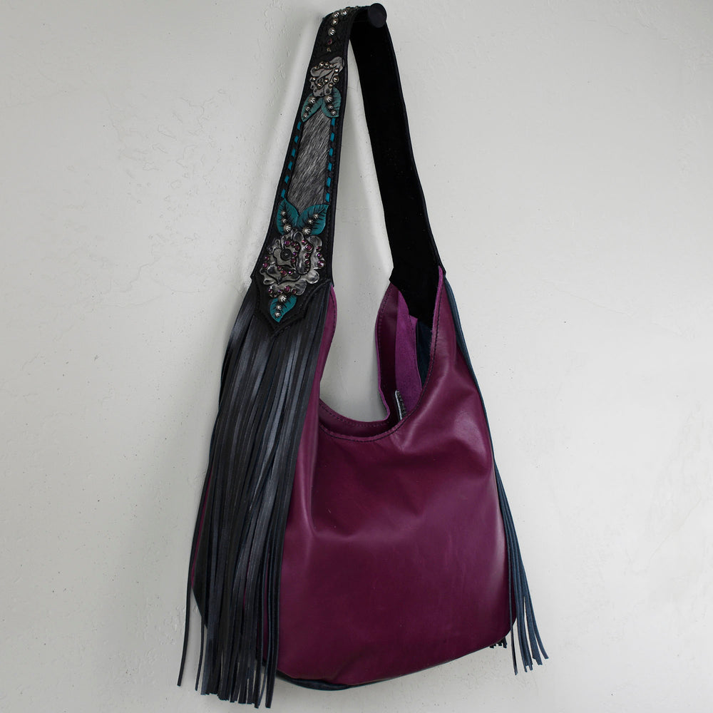 
                  
                    A purple leather Marilyn bag #2 with a black strap and decorative fringe hanging on a white wall by Heritage Brand.
                  
                