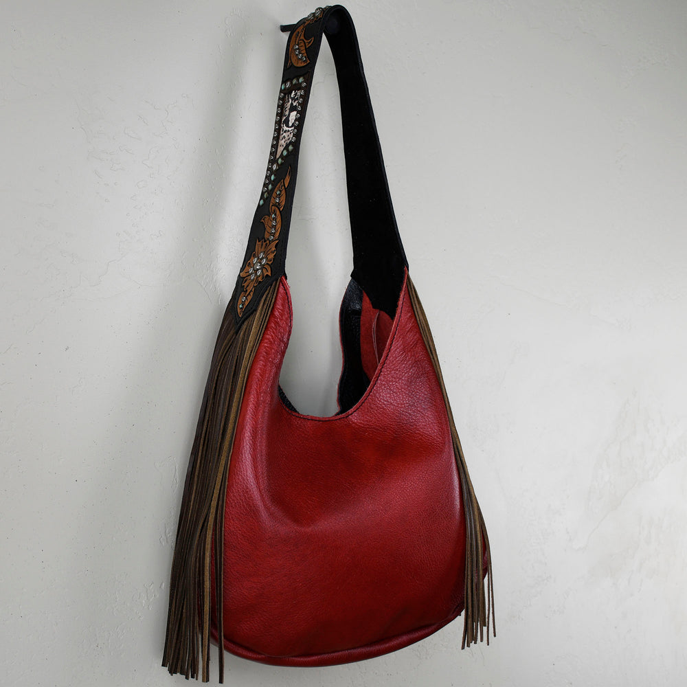
                  
                    Marilyn bag #11 by Heritage Brand, a red leather shoulder bag with a decorative strap and fringe detail, hanging against a white textured wall.
                  
                