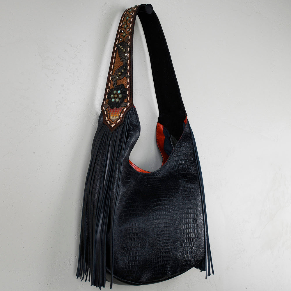 
                  
                    A black leather Marilyn bag #7 with fringe details and an embellished strap hanging against a white wall by Heritage Brand.
                  
                