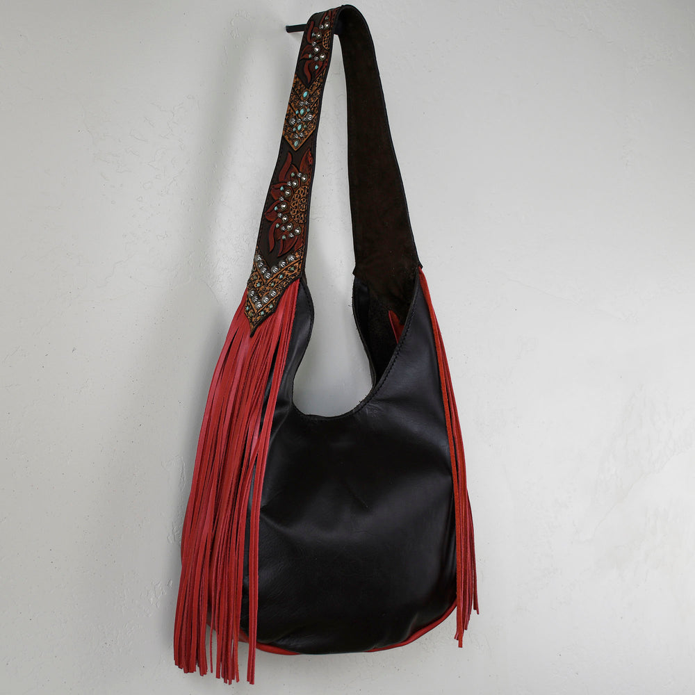 Marilyn bag #5 by Heritage Brand, a black leather shoulder bag with red fringe and decorative strap, hanging on a white wall.