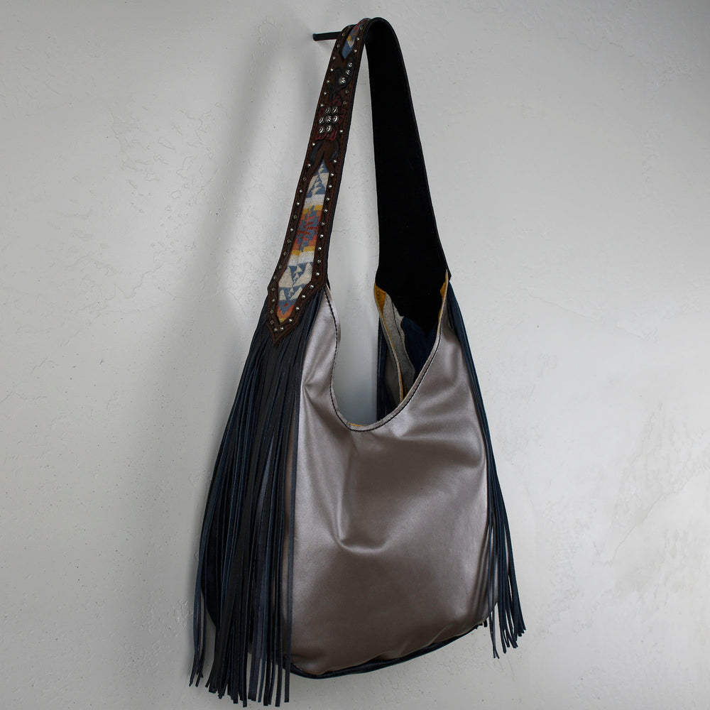 A gray Heritage Brand marilyn bag #16 with blue fringe and decorative pattern hanging against a white wall.