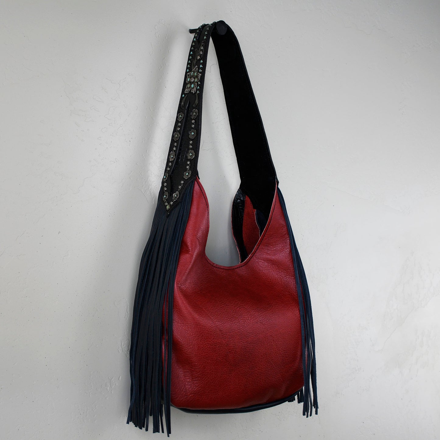 
                  
                    Marilyn bag #15 by Heritage Brand, a red leather shoulder bag with fringe and an embellished strap, hangs on a white wall.
                  
                