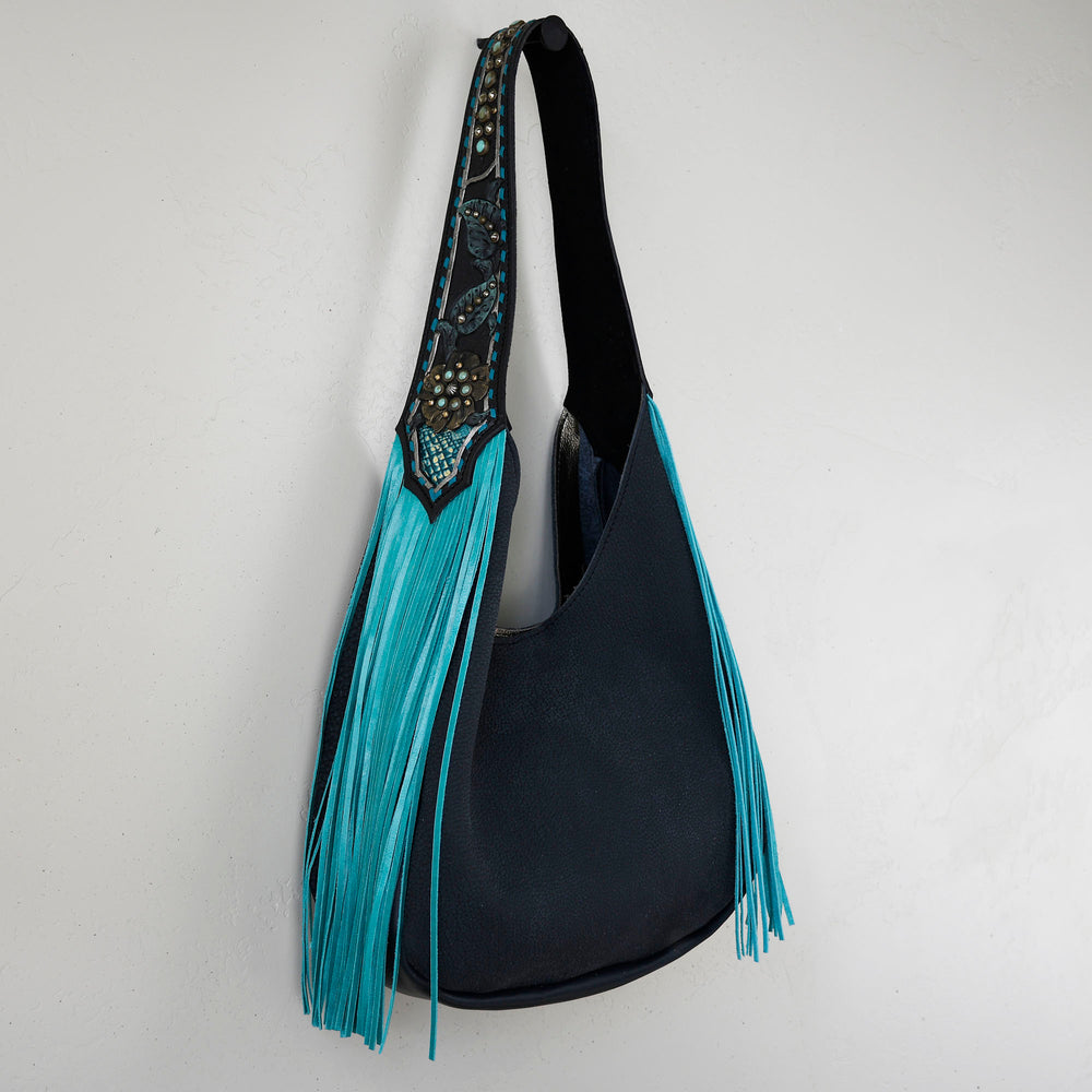 A black Marilyn bag #18 with turquoise fringe and embellishments hangs against a white wall.