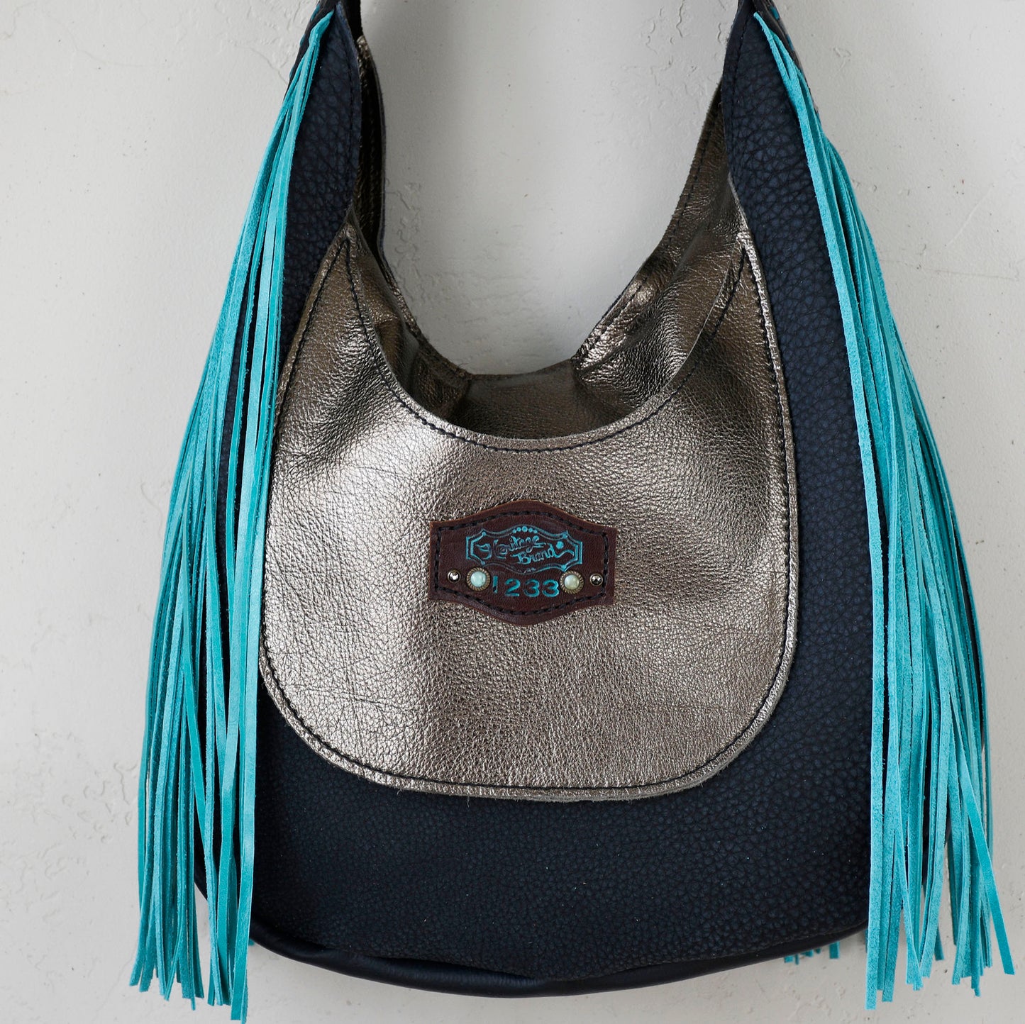 
                  
                    A silver and navy blue Marilyn Bag #18 shoulder bag with fringe detail and a Heritage Brand logo plaque.
                  
                