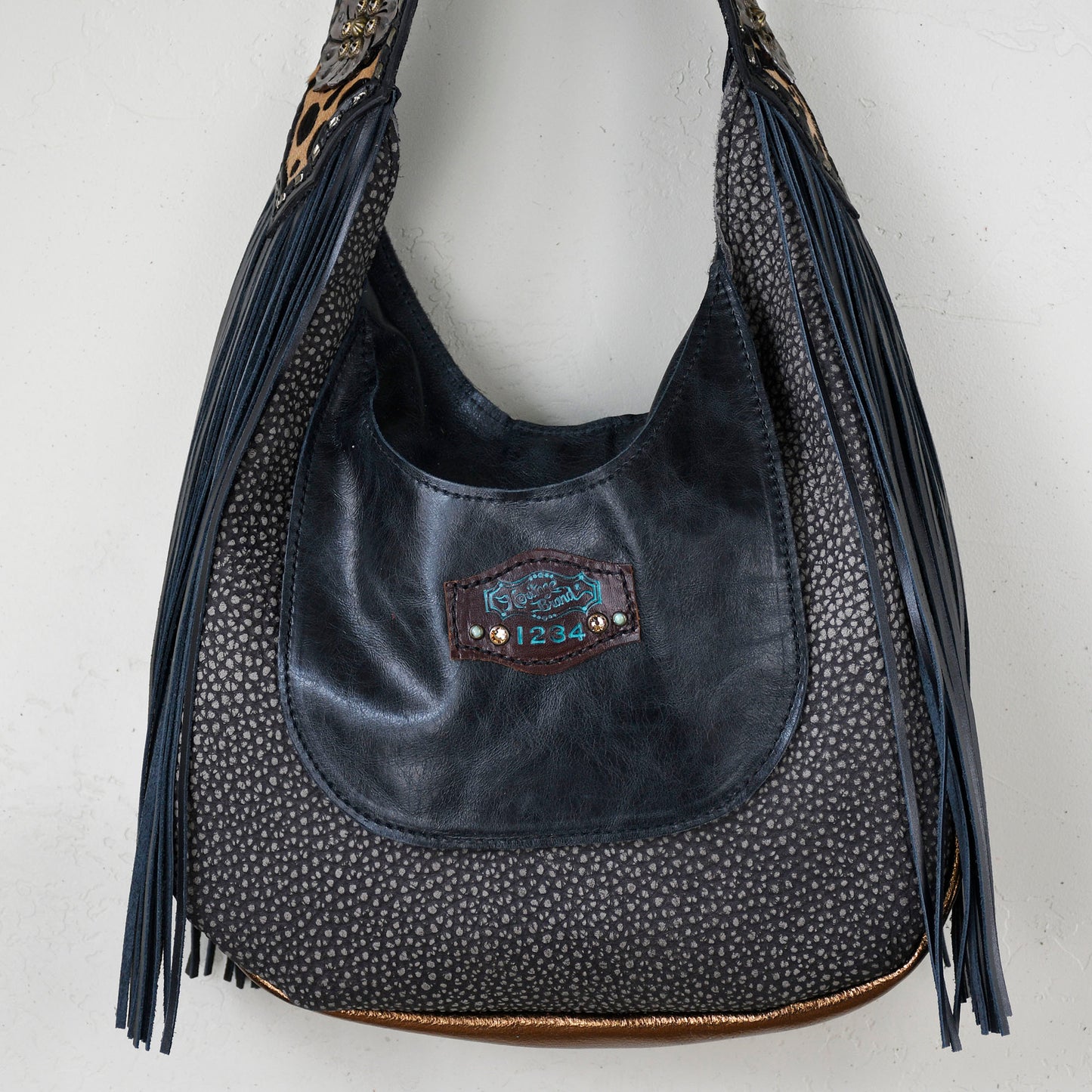 
                  
                    Marilyn bag #24 by Heritage Brand with tassels and embossed details hanging against a white wall.
                  
                