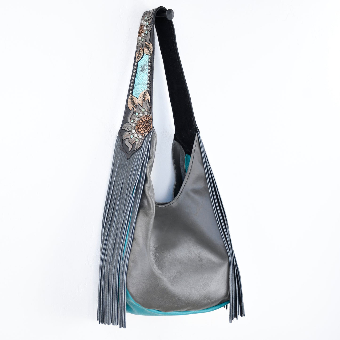 
                  
                    Heritage Brand's Marilyn Bag #1205, a gray shoulder bag with fringes and turquoise lining, stands out against a white background.
                  
                