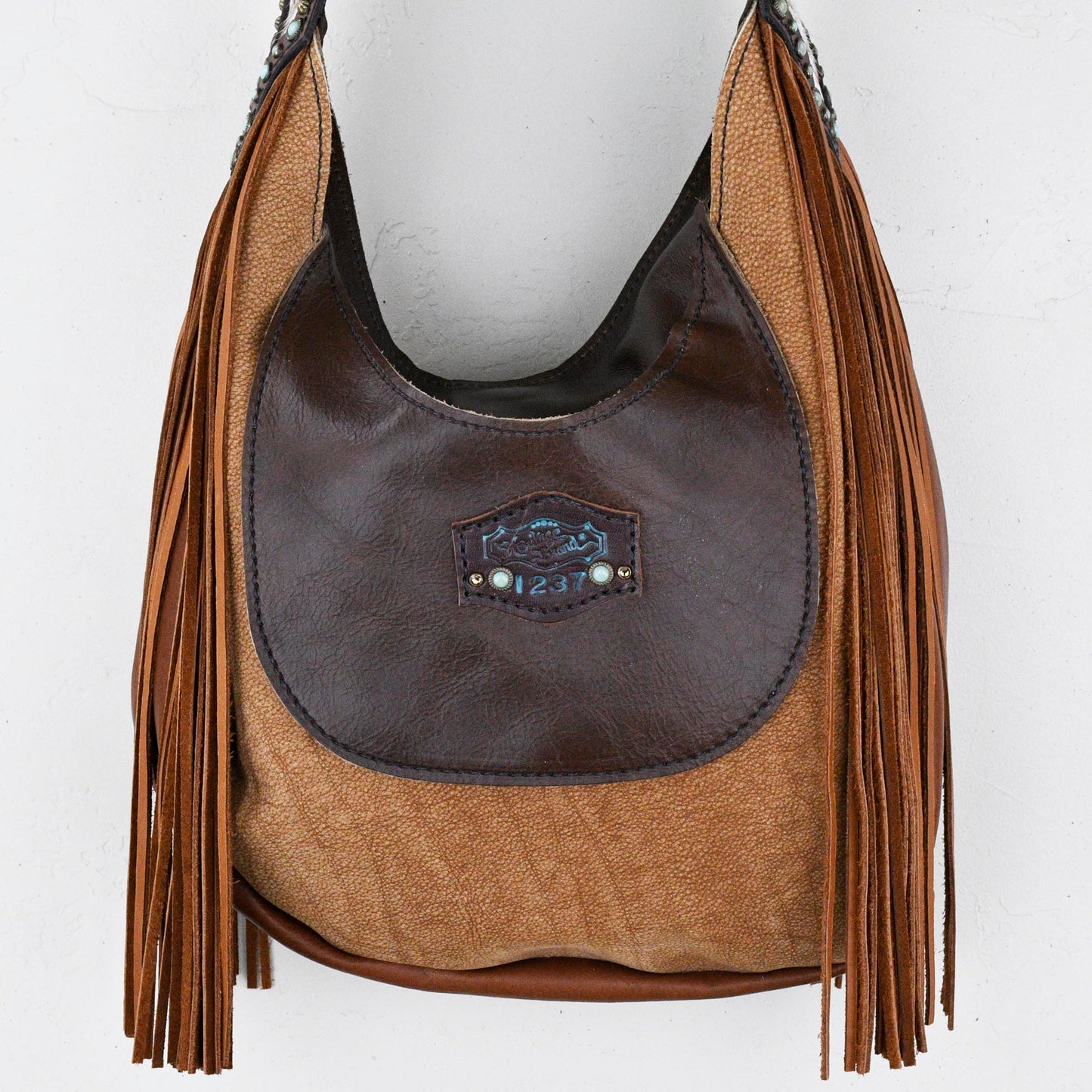 
                  
                    Brown leather and fabric Marilyn bag #28 with fringe details hanging against a white wall.
                  
                