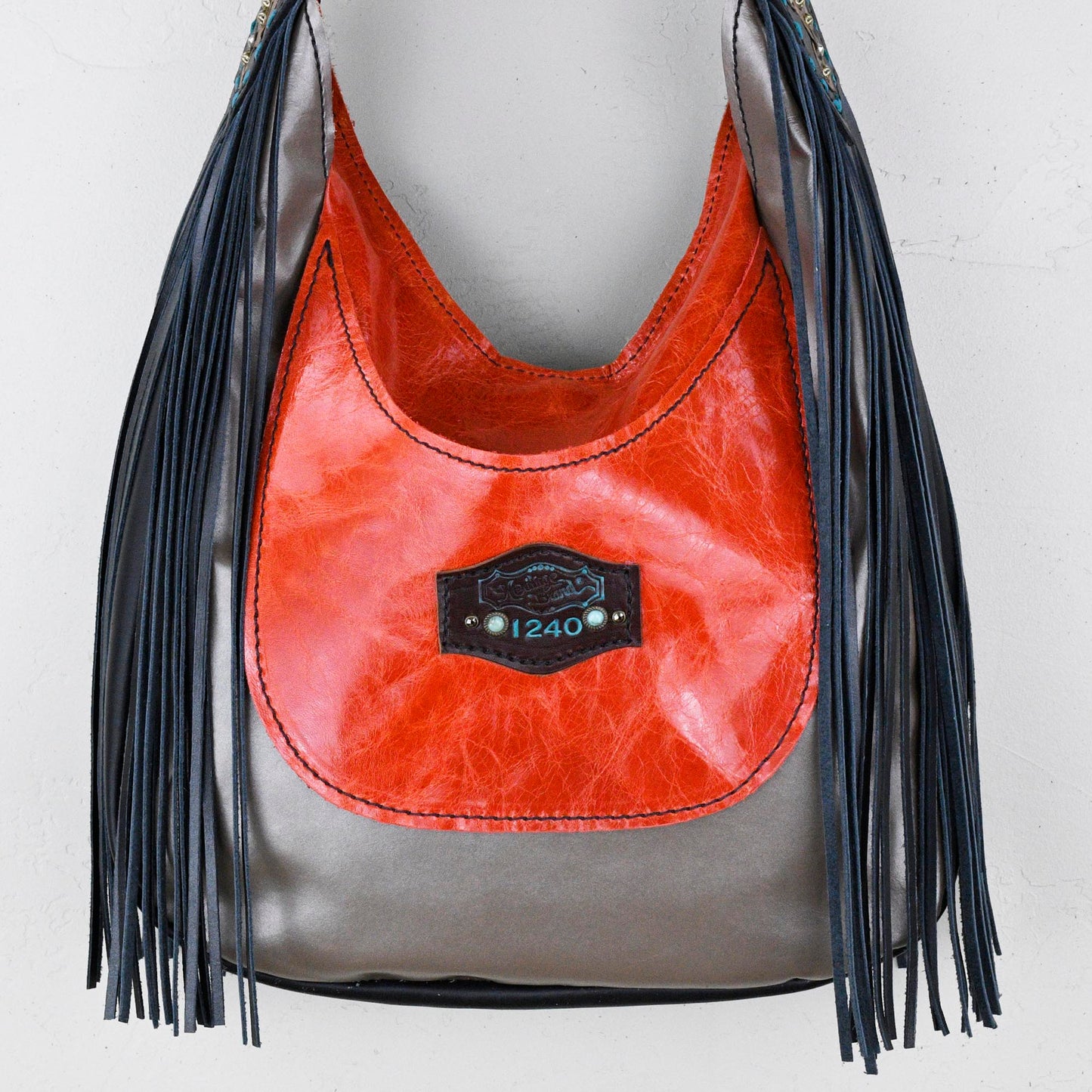 
                  
                    A red and grey leather Marilyn bag #31 with fringe detail hanging against a white wall by Heritage Brand.
                  
                