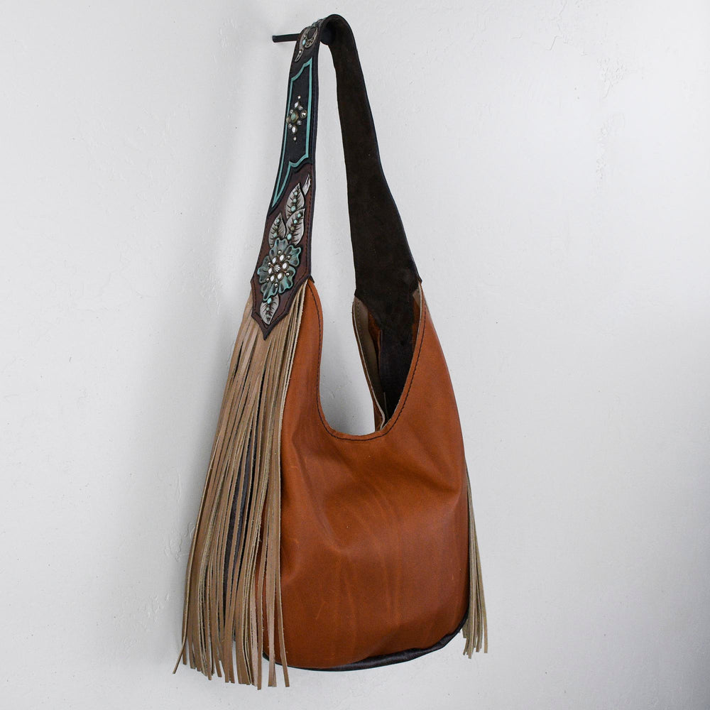 
                  
                    Marilyn bag #1242 by Heritage Brand, brown leather shoulder bag with decorative strap and fringe detail hanging against a white wall.
                  
                