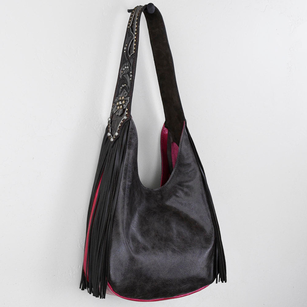 Marilyn bag #1235 by Heritage Brand, a black leather shoulder bag with fringe detail and an hand-tooled strap, is shown hanging against a white background.