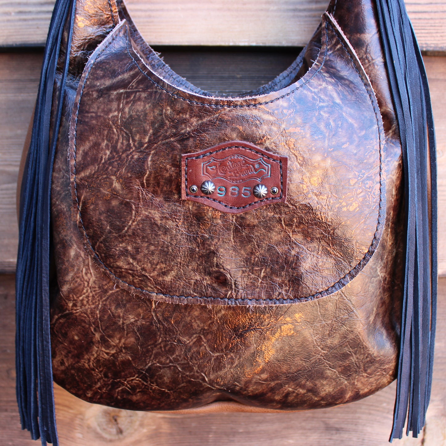 
                  
                    A brown leather crossbody marilyn bag #985 with fringe details and a metal emblem by Heritage Brand.
                  
                