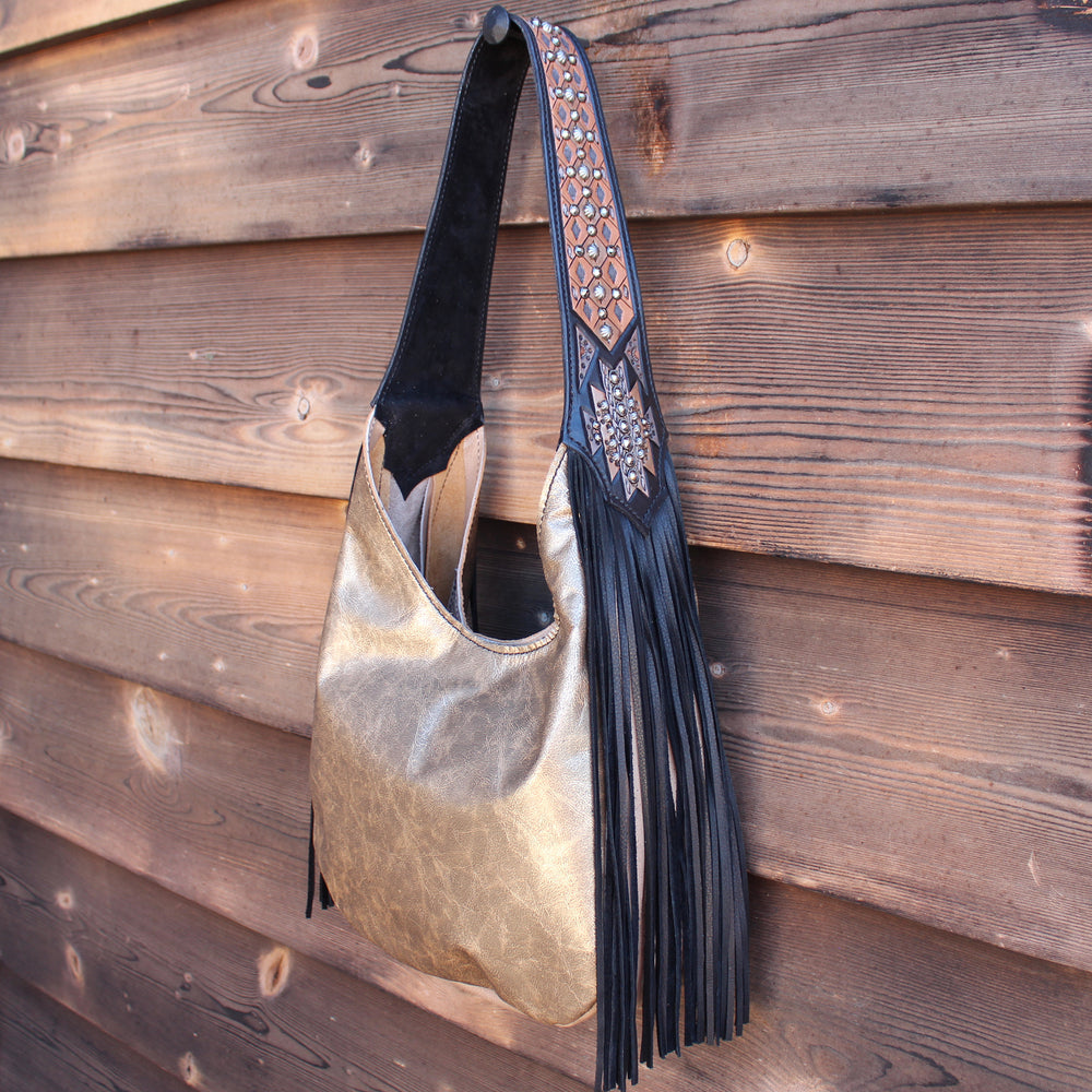 A metallic gold-colored Marilyn bag #983 with a decorative strap and tassels hanging against a wooden wall by Heritage Brand.
