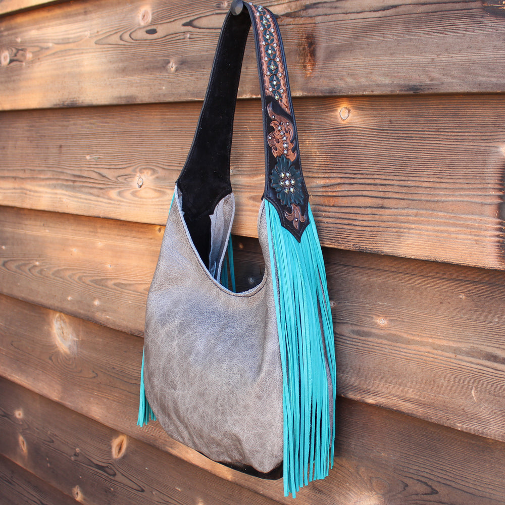 A gray suede Marilyn bag #987 with blue fringe and a decorated strap hanging against a wooden background by Heritage Brand.