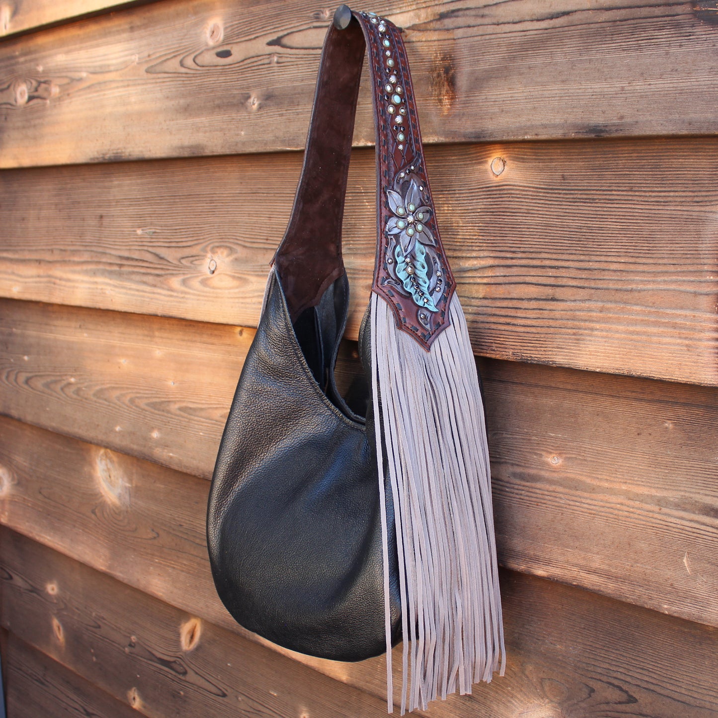 
                  
                    A black leather Heritage Brand marilyn bag #972 with decorative fringe and metallic accents hanging against a wooden backdrop.
                  
                