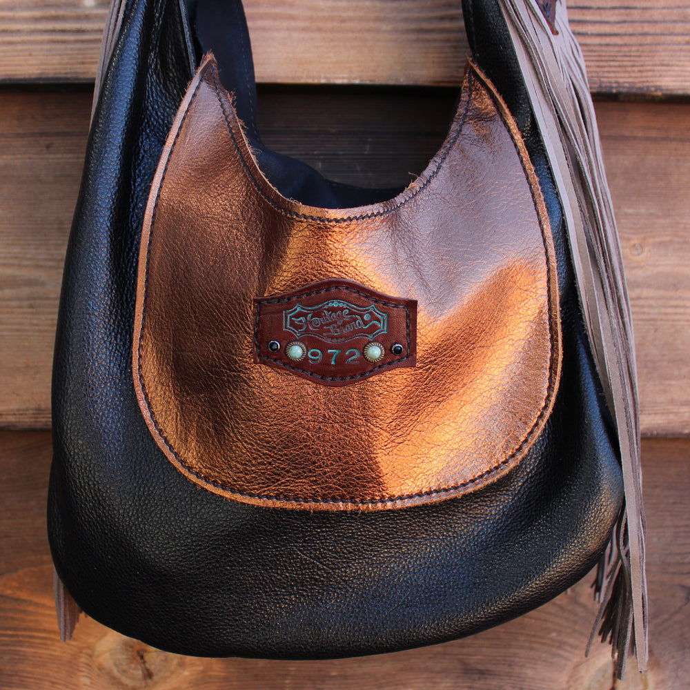 
                  
                    A brown and black leather Marilyn bag #972 with fringe detail and a Heritage Brand logo patch hanging on a wooden surface.
                  
                