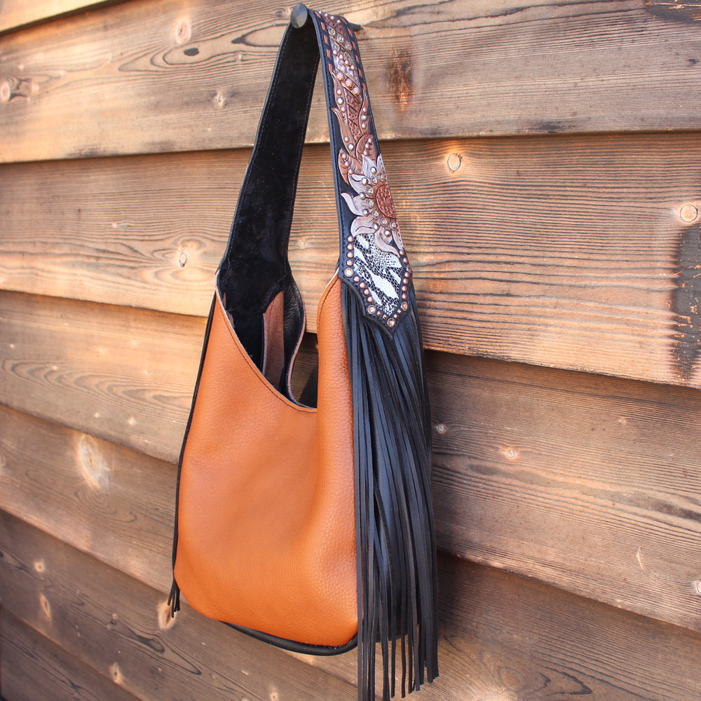 A tan-colored Marilyn bag #968 by Heritage Brand with a black strap and fringe detailing hangs on a wooden wall.