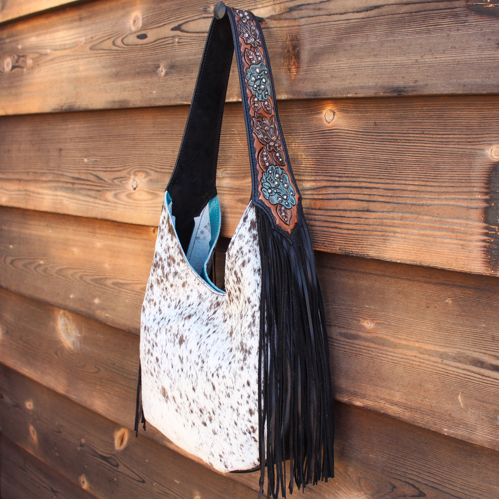 
                  
                    A stylish Marilyn Bag #984 from Heritage Brand with a cowhide pattern and fringe detailing hangs against a wooden wall.
                  
                