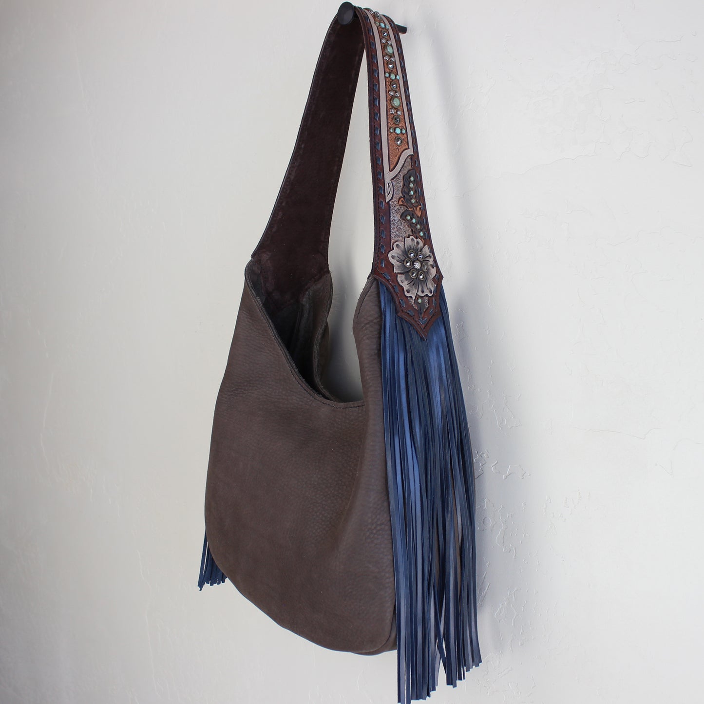 
                  
                    A brown leather Marilyn bag #1095 by Heritage Brand with a decorative strap and blue tassel hanging against a white wall.
                  
                