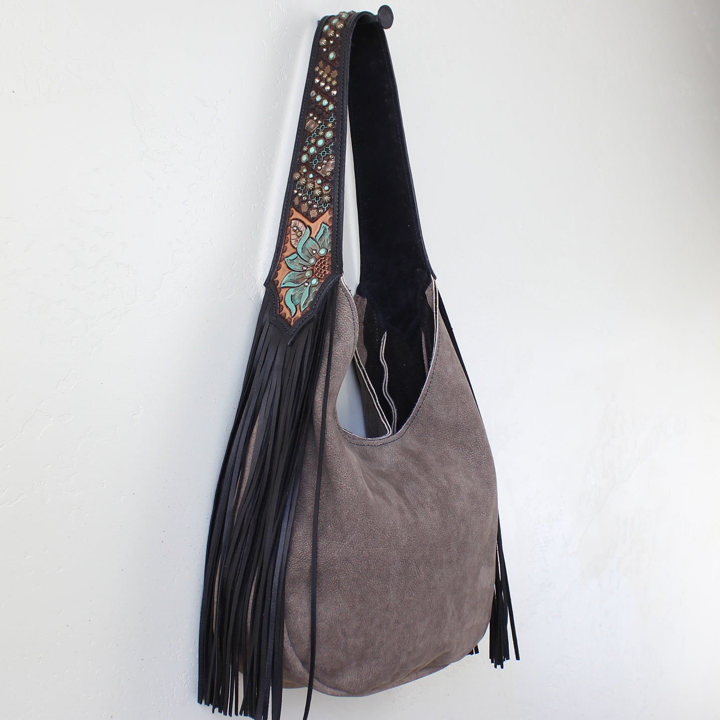 
                  
                    Gray suede Marilyn bag #1171 with decorative strap and tassel detail hanging on a white wall by Heritage Brand.
                  
                