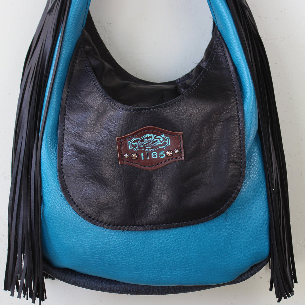
                  
                    A blue and black leather Heritage Brand marilyn bag #1185 with fringe details and a logo patch displayed on a white background.
                  
                