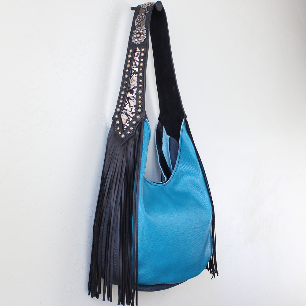 
                  
                    Blue Marilyn bag #1185 with black fringed shoulder strap hanging on a white wall by Heritage Brand.
                  
                