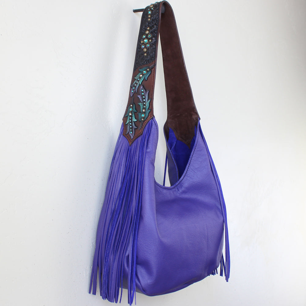 A purple leather fringed marilyn bag #1186 with a decorative strap hanging on a white wall by Heritage Brand.