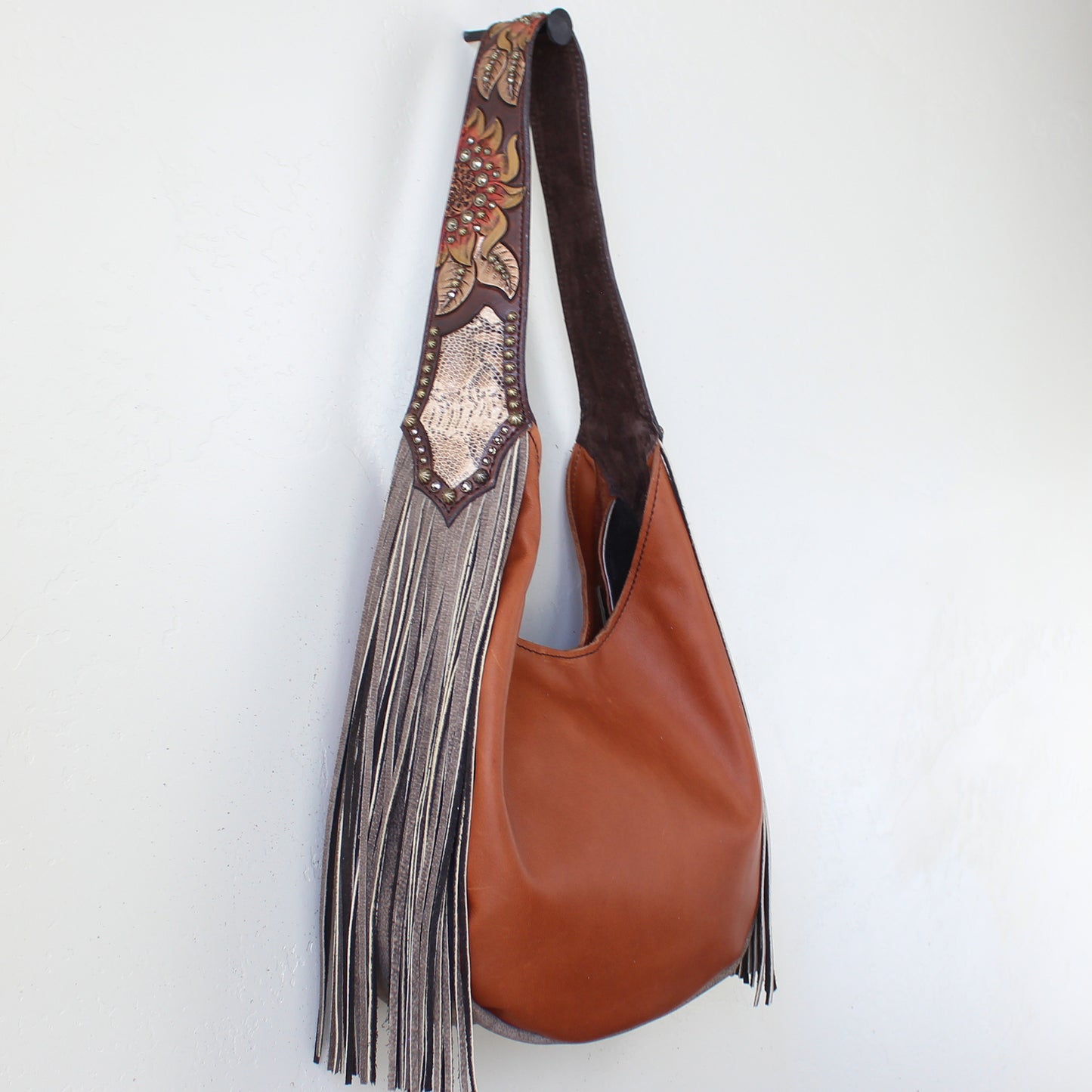 
                  
                    Marilyn bag #1189 by Heritage Brand with decorative fringe and hand-tooled strap hanging against a white wall.
                  
                