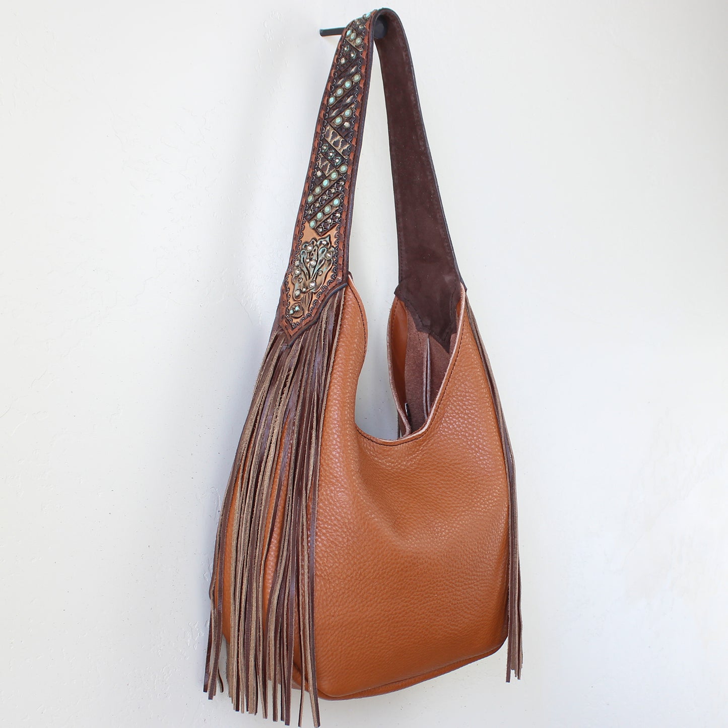 
                  
                    Brown leather marilyn bag #1191 by Heritage Brand with decorative fringe and embossed strap hanging against a white wall.
                  
                