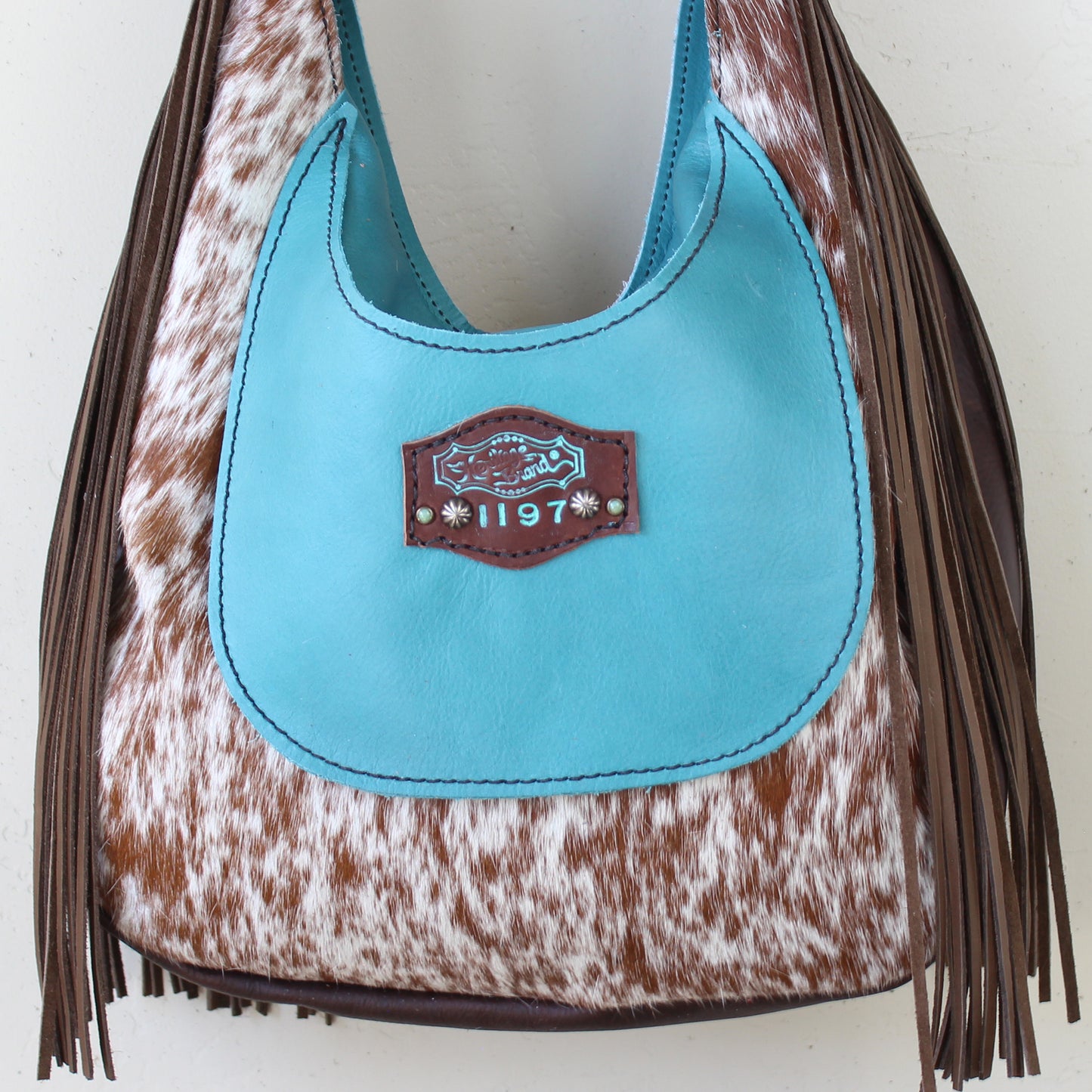 
                  
                    A blue and brown fringed shoulder bag with cowhide pattern and a metal emblem, the marilyn bag #1197 by Heritage Brand.
                  
                