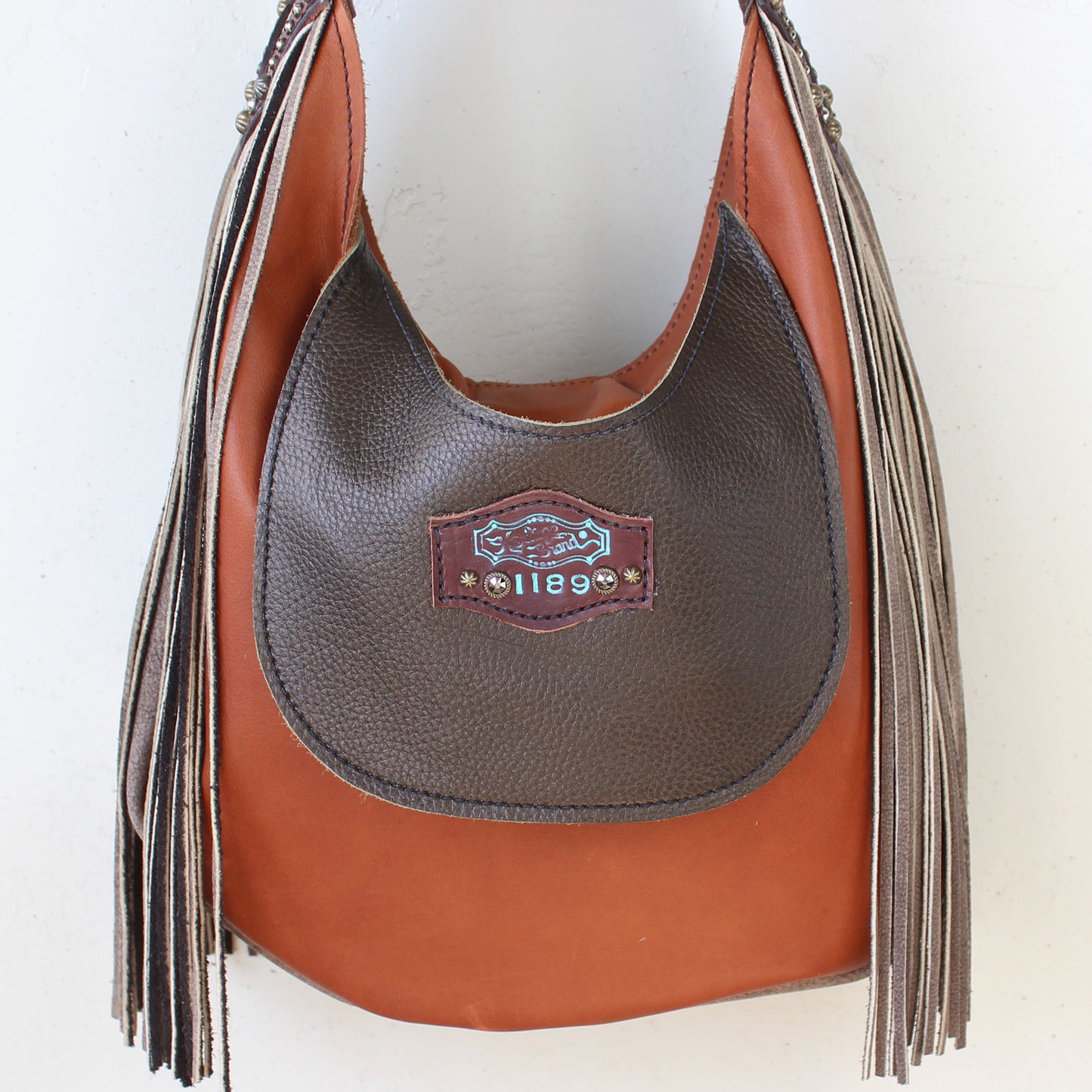 
                  
                    Brown and tan leather Heritage Brand marilyn bag #1189 with fringe details and a brand logo on the front.
                  
                